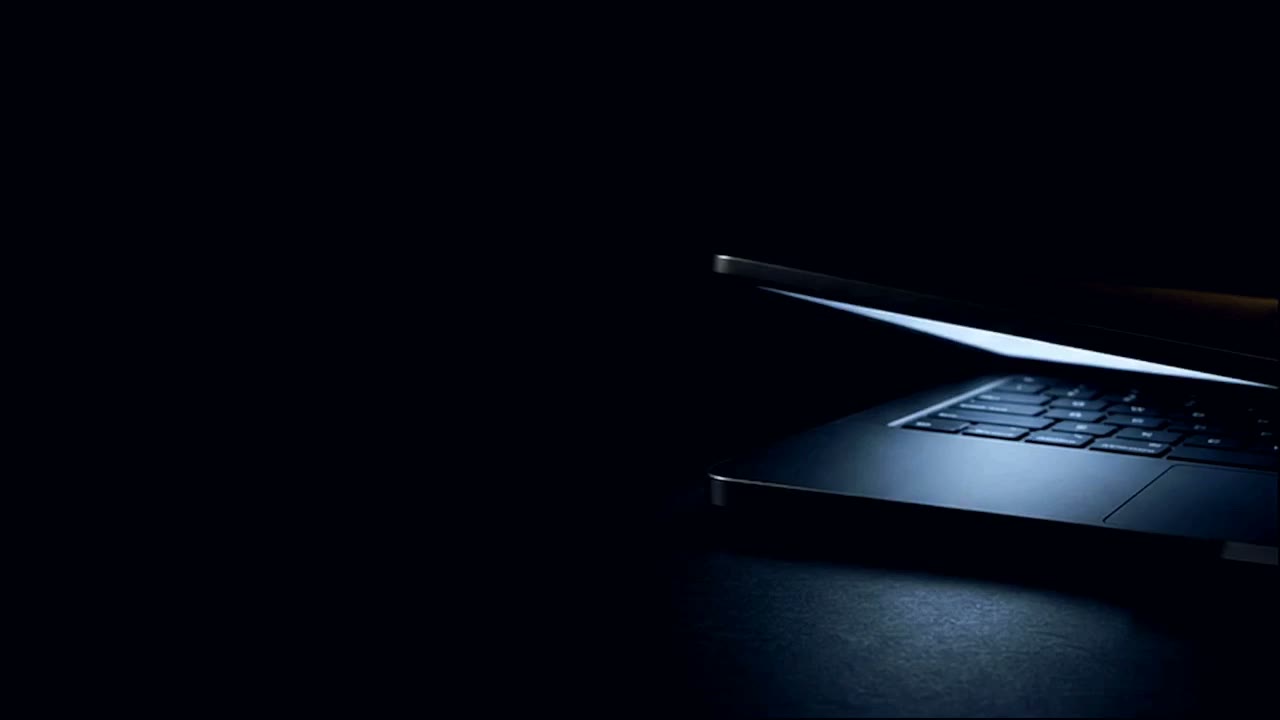 A MacBook in the dark with its lid almost closed, the cool light of its still-lit screen spilling out of the narrowing gap and across the desk.